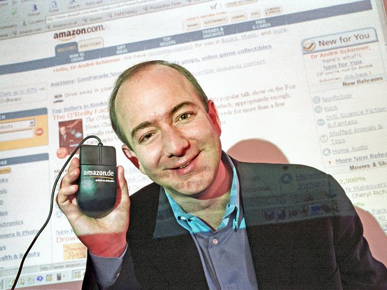 Jeff Bezos im Jahr 2000. Er lächelt in die Kamera und hält eine Computermaus nach oben, auf der Amazon steht. Im Hintergrund ist auf einer Leinwand die Amazon-Webseite zu sehen.