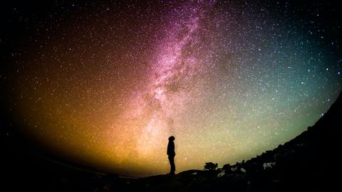 Unter einem bunten Sternenhimmel ist die Silhouette einer Person, die nach oben schaut, zu erkennen.