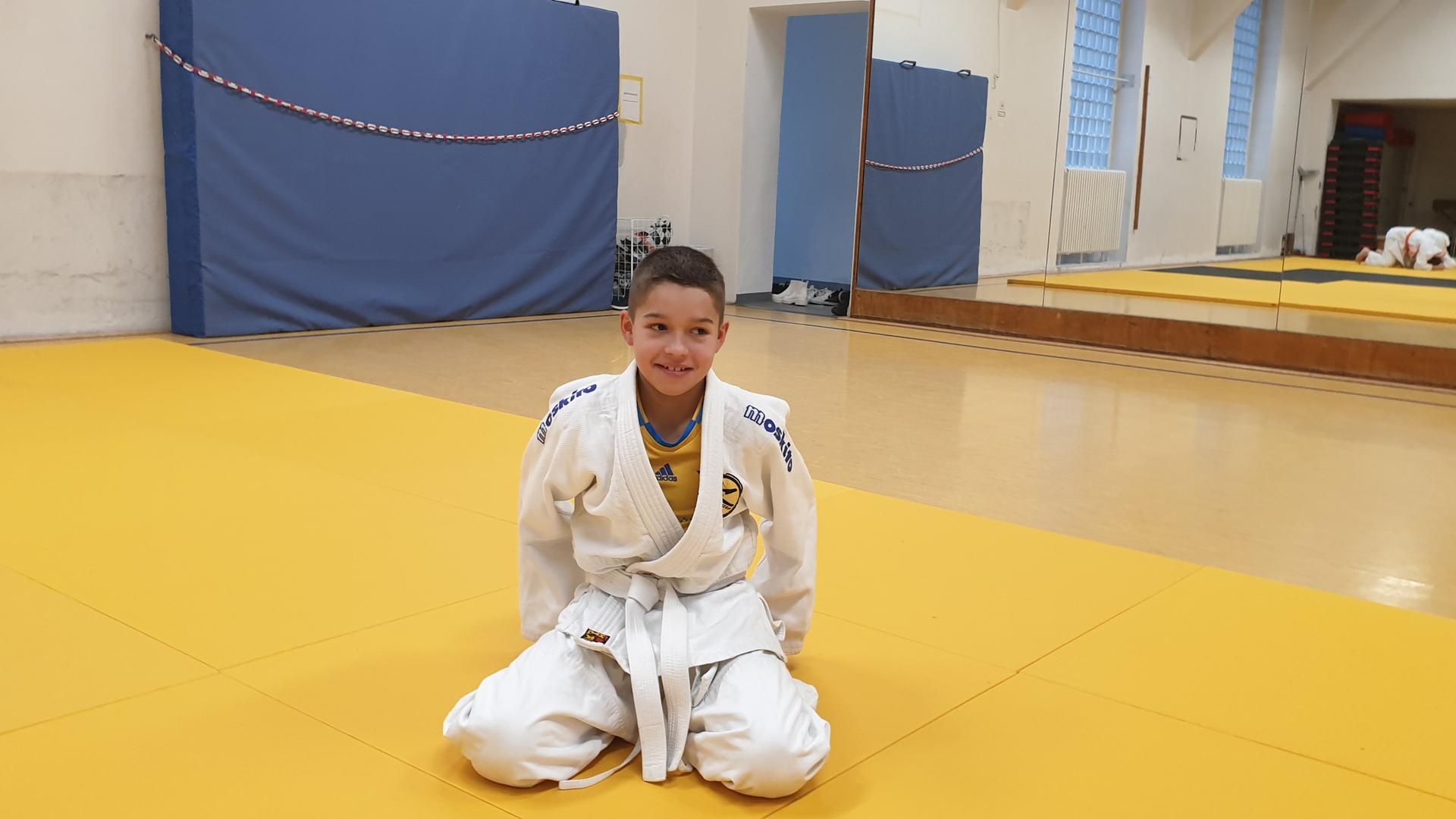 Der elfjährige Pavlo sitzt mit einem Judoanzug, einem Keikogi, bekleidet auf einer Judomatte in einer Turnhalle.