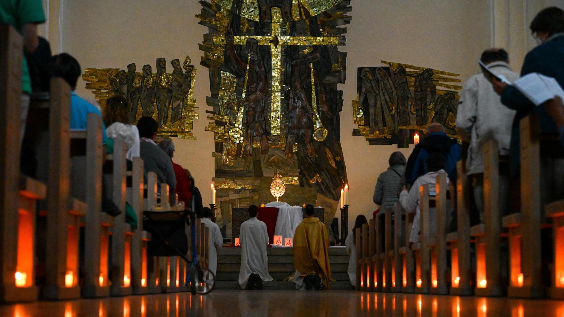 Brennende Kerzen sind auf dem Boden unter den Kirchenbänken aufgestellt, auf denen Teilnehmer sitzen. Am Altar knien Menschen in kirchlichen Roben.