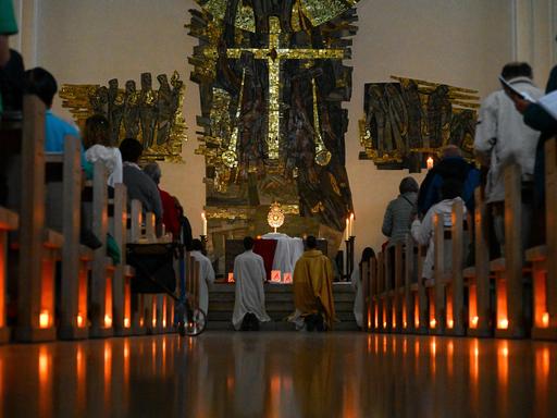 Brennende Kerzen sind auf dem Boden unter den Kirchenbänken aufgestellt, auf denen Teilnehmer sitzen. Am Altar knien Menschen in kirchlichen Roben. Brennende Kerzen sind auf dem Boden unter den Kirchenbänken aufgestellt, auf denen Teilnehmer sitzen. Am Altar knien Menschen in kirchlichen Roben.