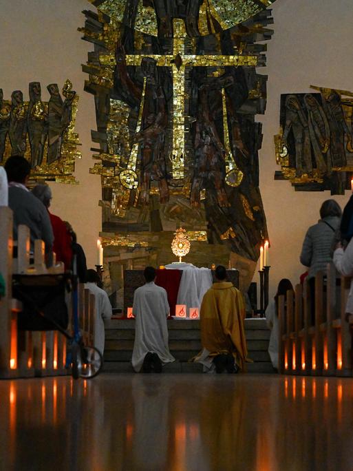 Brennende Kerzen sind auf dem Boden unter den Kirchenbänken aufgestellt, auf denen Teilnehmer sitzen. Am Altar knien Menschen in kirchlichen Roben. Brennende Kerzen sind auf dem Boden unter den Kirchenbänken aufgestellt, auf denen Teilnehmer sitzen. Am Altar knien Menschen in kirchlichen Roben.