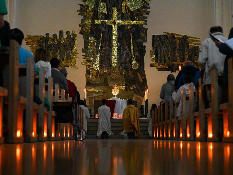 Brennende Kerzen sind auf dem Boden unter den Kirchenbänken aufgestellt, auf denen Teilnehmer sitzen. Am Altar knien Menschen in kirchlichen Roben.