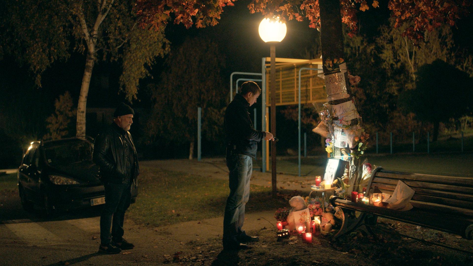 Zwei Männer stehen im Dunkeln am Tatort eines Mordfalls. Dort brennen Kerzen, die an das Opfer erinnern.
