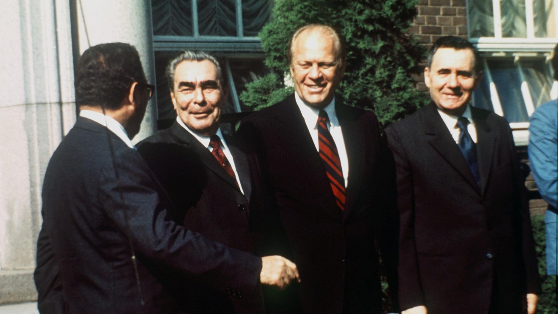 Ein Farbfoto zeigt vier lachende Männer. In der Bildmitte reichen sich der sowjetische Parteichef Leonid Breschnew, und US-Präsident Gerald Ford die Hand.