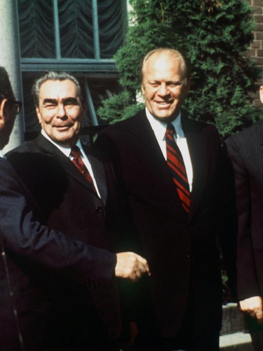 Ein Farbfoto zeigt vier lachende Männer. In der Bildmitte reichen sich der sowjetische Parteichef Leonid Breschnew, und US-Präsident Gerald Ford die Hand.