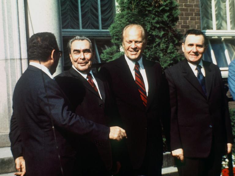 Ein Farbfoto zeigt vier lachende Männer. In der Bildmitte reichen sich der sowjetische Parteichef Leonid Breschnew, und US-Präsident Gerald Ford die Hand.