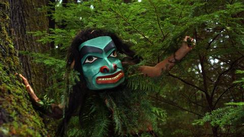 Staas Guujaaw bei einem Tanz auf dem Golden Spruce Trail auf der Insel Queen Charlotte in British Columbia, Kanada. Die Person trägt eine türkis-grüne Maske in grünen Büschen.