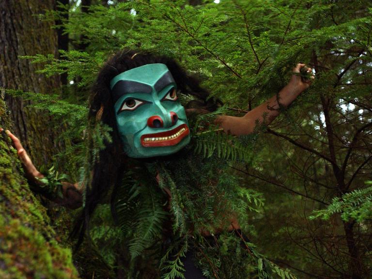 Staas Guujaaw bei einem Tanz auf dem Golden Spruce Trail auf der Insel Queen Charlotte in British Columbia, Kanada. Die Person trägt eine türkis-grüne Maske in grünen Büschen.