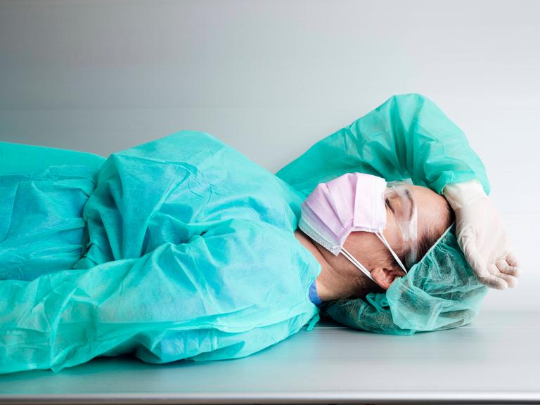 Eine Ärztin liegt in Schutzkleidung erschöpft auf einer Metallbank.