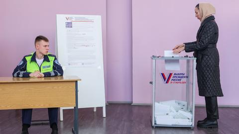 Eine Frau wirft in Rjasan un Russland ihren Stimmzettel in eine Wahlurne. Daneben sitzt ein Wahlhelfer in Uniform und gelber Weste an einem Tisch.