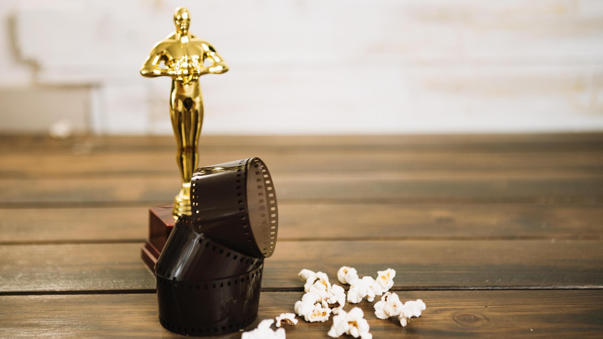 Das Symbolbild zeigt eine Oscar-Statuette, eine Filmrolle und Popcorn auf Holzdielen.