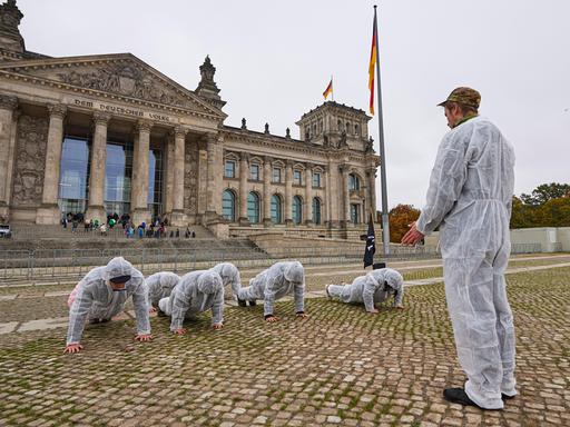 Eine Gruppe von jungen Menschen in weißen Schutzanzügen bei einer Protestaktion vor dem Bundestag. Eine Person steht und trägt eine Camouflage Kopfbedeckung, die anderen machen Liegestütze. Die Situation erinnert an Trainings bei der Bundeswehr.