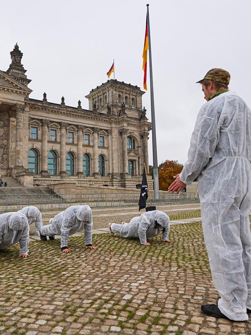 Eine Gruppe von jungen Menschen in weißen Schutzanzügen bei einer Protestaktion vor dem Bundestag. Eine Person steht und trägt eine Camouflage Kopfbedeckung, die anderen machen Liegestütze. Die Situation erinnert an Trainings bei der Bundeswehr.
