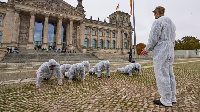 Eine Gruppe von jungen Menschen in weißen Schutzanzügen bei einer Protestaktion vor dem Bundestag. Eine Person steht und trägt eine Camouflage Kopfbedeckung, die anderen machen Liegestütze. Die Situation erinnert an Trainings bei der Bundeswehr.