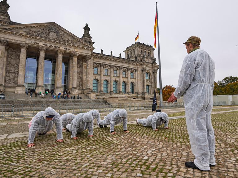 Eine Gruppe von jungen Menschen in weißen Schutzanzügen bei einer Protestaktion vor dem Bundestag. Eine Person steht und trägt eine Camouflage Kopfbedeckung, die anderen machen Liegestütze. Die Situation erinnert an Trainings bei der Bundeswehr.