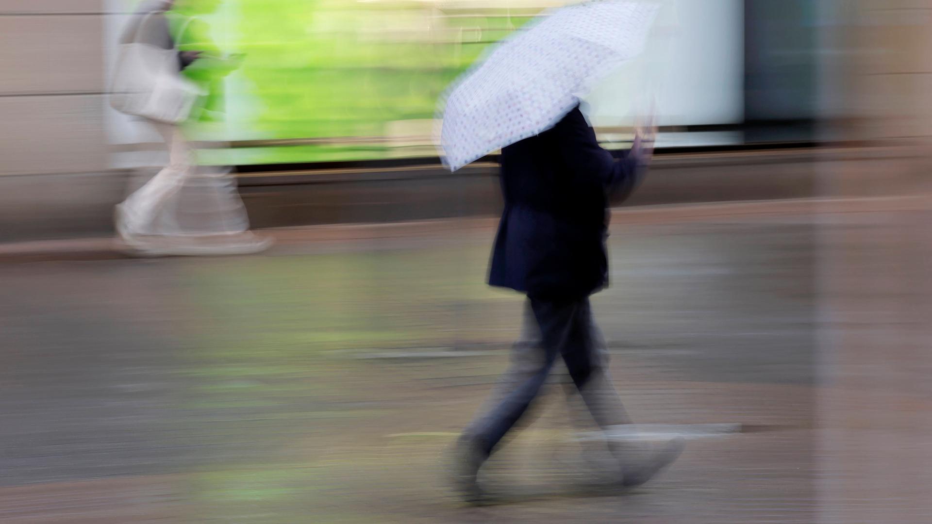 Passanten laufen im Nieselregen mit Schirmen durch eine Fußgängerzone. Die Aufnahme ist verzerrt. Es ist ein Symbolbild für die Erkältungszeit.