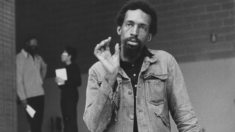 Auf dem schwarz-weißen Bild steht ein farbiger Mann in Jeansjacke und gestikuliert mit der rechten Hand, während er sich mit der anderen Hand aufstützt. 