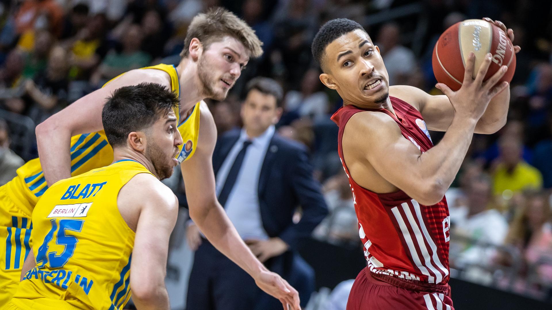 Jason George verteidigt den Ball. Er trägt ein rotes Trikot von Bayern München. Zwei Spieler von Alba Berlin kämpfen um den Ball, sie tragen gelbe Trikots.