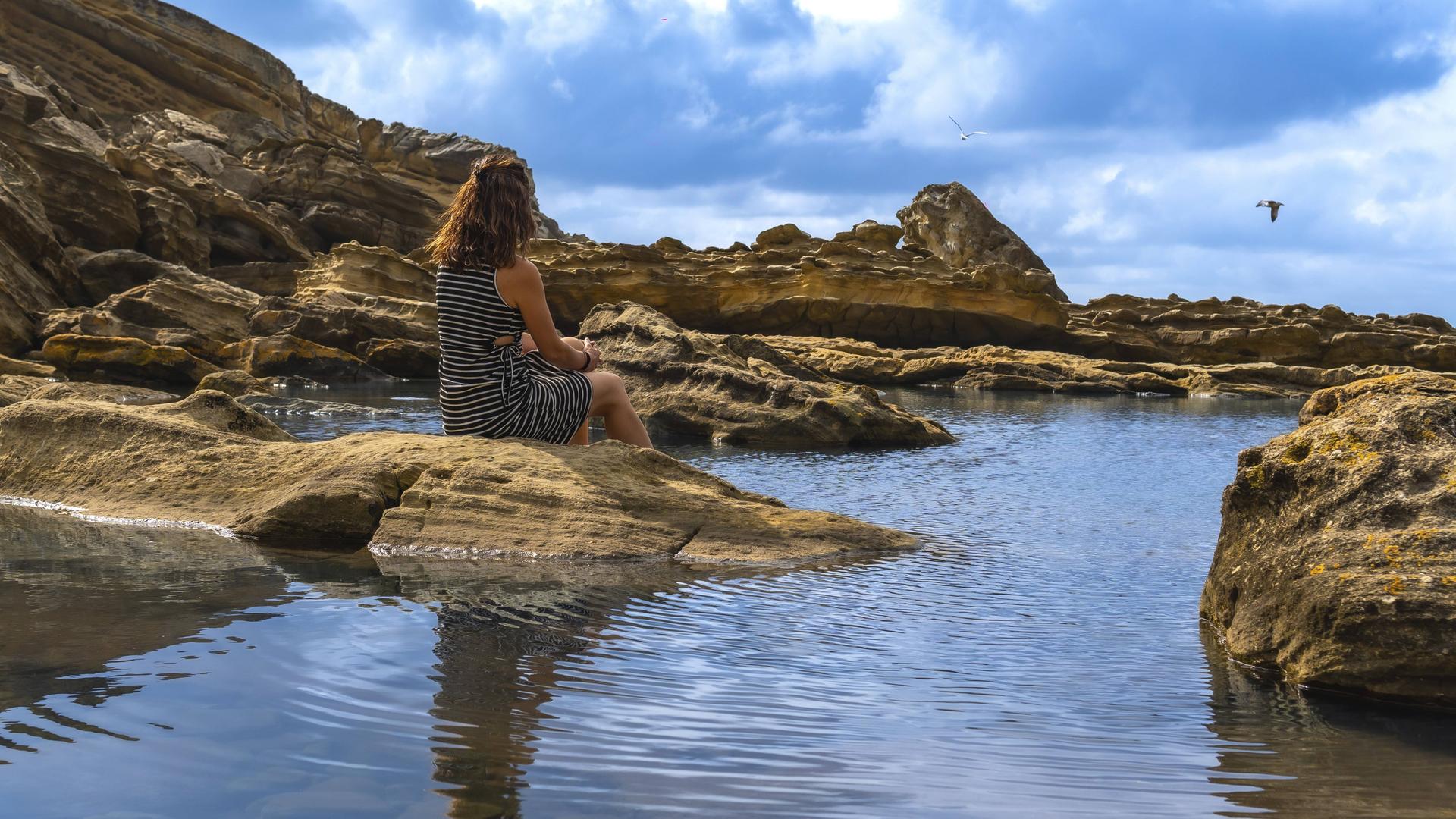 Frau sitzt auf alten Felsen an einem ruhigen Flutbecken, beobachtet das ruhige Meer und die zerklüftete Küste des Mount Jaizkibel im Baskenland und findet Einsamkeit in der Natur. 