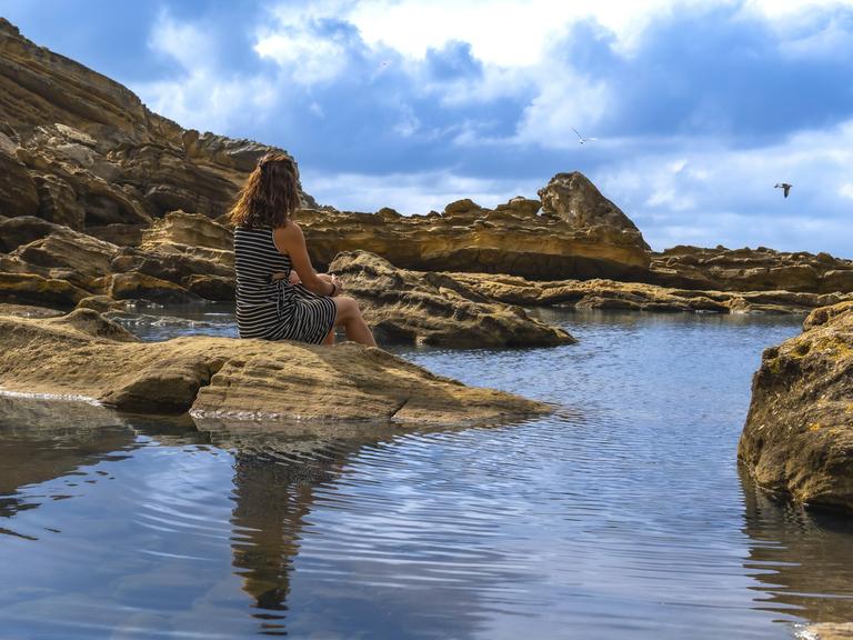 Frau sitzt auf alten Felsen an einem ruhigen Flutbecken, beobachtet das ruhige Meer und die zerklüftete Küste des Mount Jaizkibel im Baskenland und findet Einsamkeit in der Natur. 