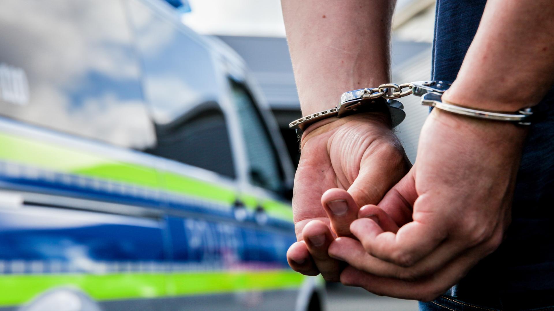 Eine Person steht in Hand-Schellen gefesselt neben einem Polizei-Auto. Eine Person steht in Hand-Schellen gefesselt neben einem Polizei-Auto.