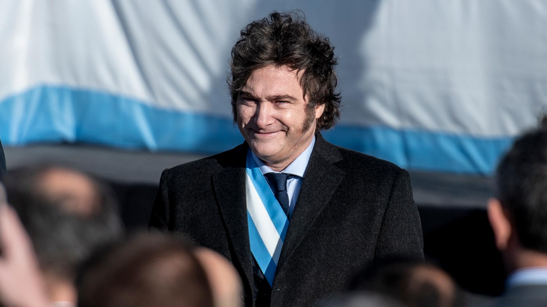 Javier Milei trägt eine blau-weiße Schärpe unter seinem schwarzen Sakko, steht vor der argentinischen Flagge und lächelt ins Publikum