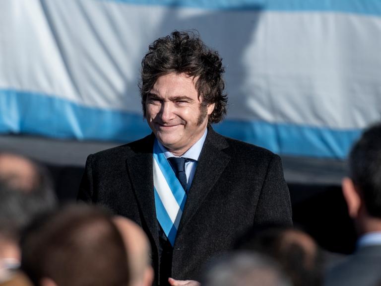 Javier Milei trägt eine blau-weiße Schärpe unter seinem schwarzen Sakko, steht vor der argentinischen Flagge und lächelt ins Publikum