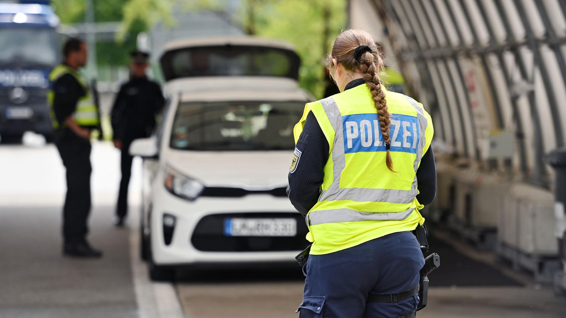 Grenzpolizisten an der Grenze zu Österreich. Im Vordergrund ist eine Polizistin von hinten zu sehen, im Hintergrund zwei Polizisten bei der Kontrolle eines Autos. Grenzpolizisten an der Grenze zu Österreich. Im Vordergrund ist eine Polizistin von hinten zu sehen, im Hintergrund zwei Polizisten bei der Kontrolle eines Autos.