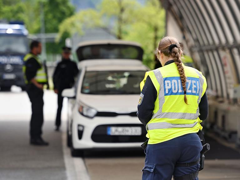 Grenzpolizisten an der Grenze zu Österreich. Im Vordergrund ist eine Polizistin von hinten zu sehen, im Hintergrund zwei Polizisten bei der Kontrolle eines Autos.