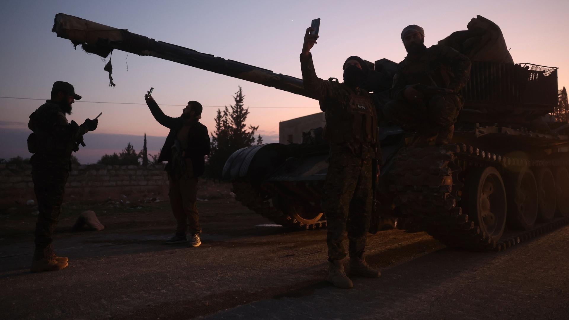 Im Abendrot stehen mehrere Männner vor einem Panzer und machen Selfies mit Mobiltelefonen. Sie sind als Silhouetten zu sehen. 