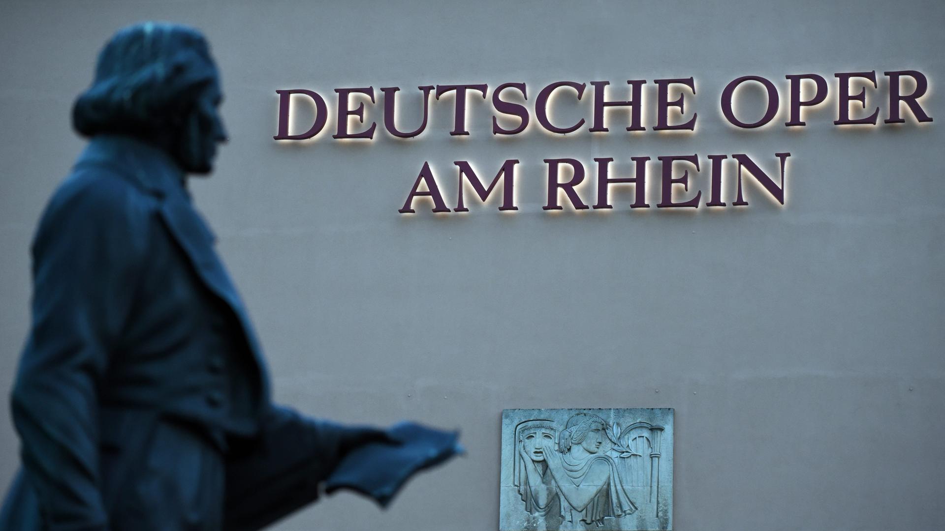 Das beleuchtete Logo "Deutsche Oper am Rhein" am Gebäude der Deutschen Oper am Rhein in Düsseldorf. Im Vordergrund die unscharfe Statue des Komponisten Felix Mendelssohn Bartholdy