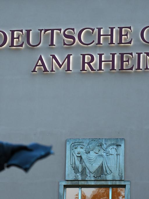Das beleuchtete Logo "Deutsche Oper am Rhein" am Gebäude der Deutschen Oper am Rhein in Düsseldorf. Im Vordergrund die unscharfe Statue des Komponisten Felix Mendelssohn Bartholdy.