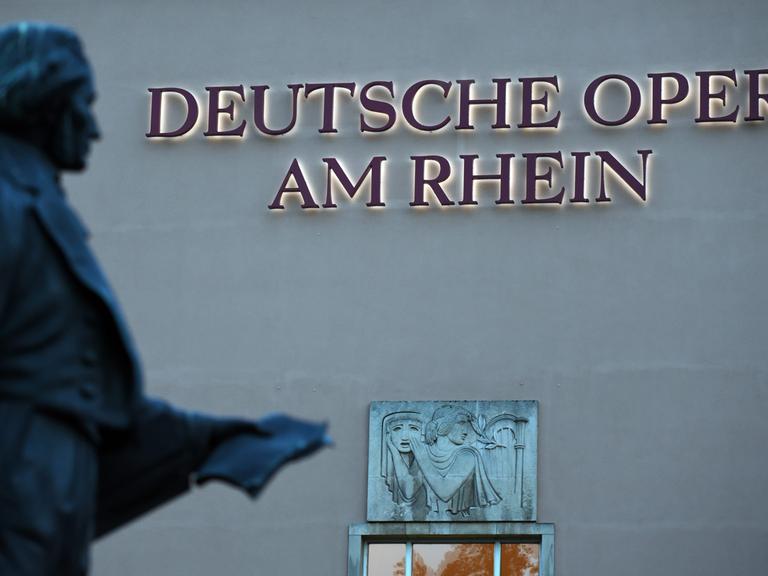 Das beleuchtete Logo "Deutsche Oper am Rhein" am Gebäude der Deutschen Oper am Rhein in Düsseldorf. Im Vordergrund die unscharfe Statue des Komponisten Felix Mendelssohn Bartholdy.