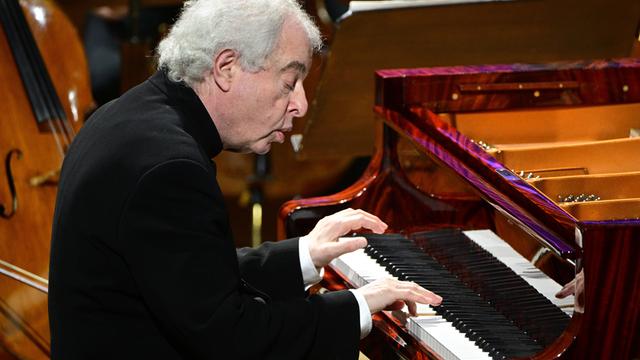 Der Pianist András Schiff sitzt an einem schwarzen Flügel und spielt. Er sitzt leicht nach vorne gebeugt und schaut sehr konzentriert. 

