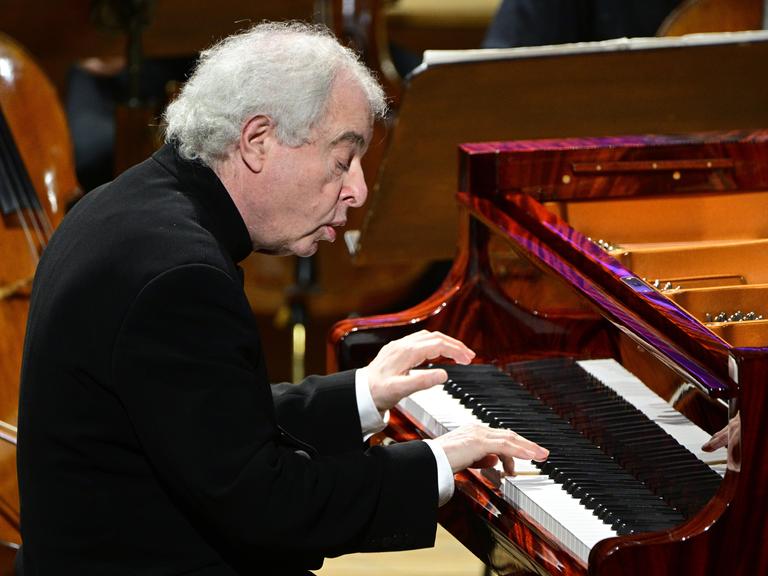 Der Pianist András Schiff sitzt an einem schwarzen Flügel und spielt. Er sitzt leicht nach vorne gebeugt und schaut sehr konzentriert. 
