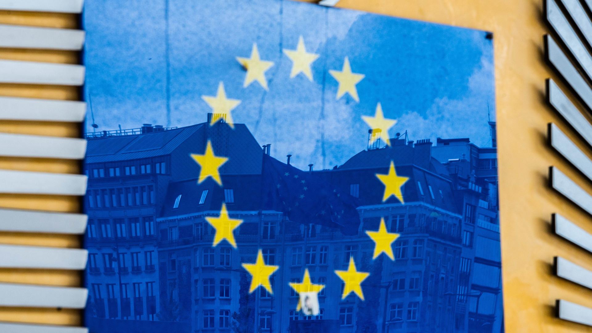 Drei Fahnen der Europäischen Union und gegenüberliegende Gebäude spiegeln sich im EU-Logo an der Außenfassade des Berlaymont-Gebäudes in Brüssel. Drei Fahnen der Europäischen Union und gegenüberliegende Gebäude spiegeln sich im EU-Logo an der Außenfassade des Berlaymont-Gebäudes in Brüssel.