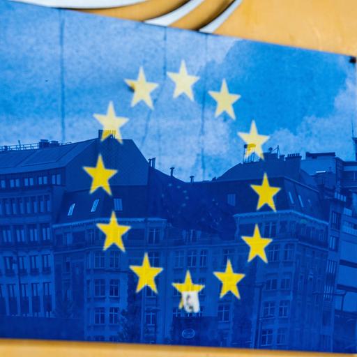 Drei Fahnen der Europäischen Union und gegenüberliegende Gebäude spiegeln sich im EU-Logo an der Außenfassade des Berlaymont-Gebäudes in Brüssel.