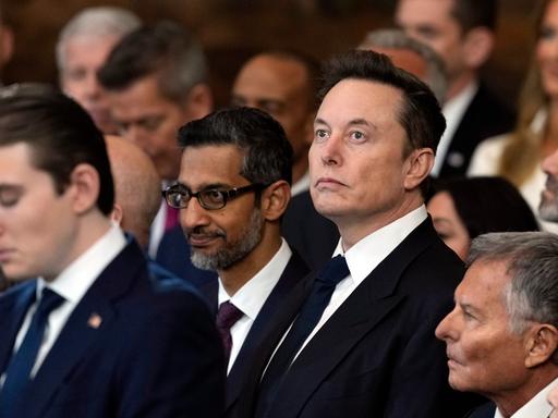 Elon Musk steht inmitten von Menschen im Capitol bei der Amtseinführung von Donald Trump und schaut nach oben.