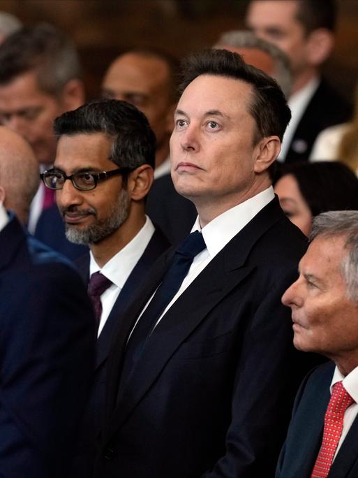 Elon Musk steht inmitten von Menschen im Capitol bei der Amtseinführung von Donald Trump und schaut nach oben.