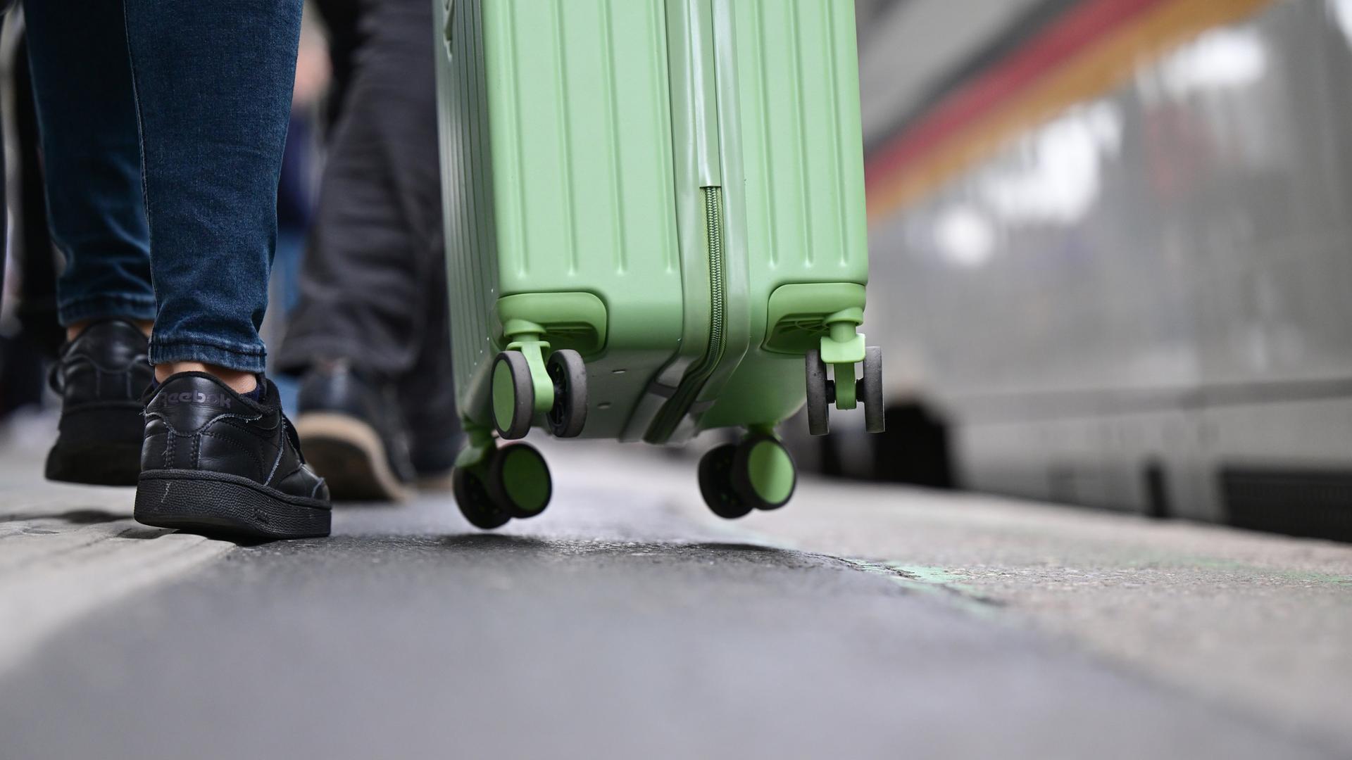 Ein hellgrüner Rollkoffer wird über einen Bahnsteig gezogen.