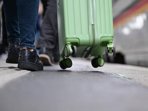 Ein hellgrüner Rollkoffer wird über einen Bahnsteig gezogen, an der Wand ein schwarz-rot-goldenes Band.