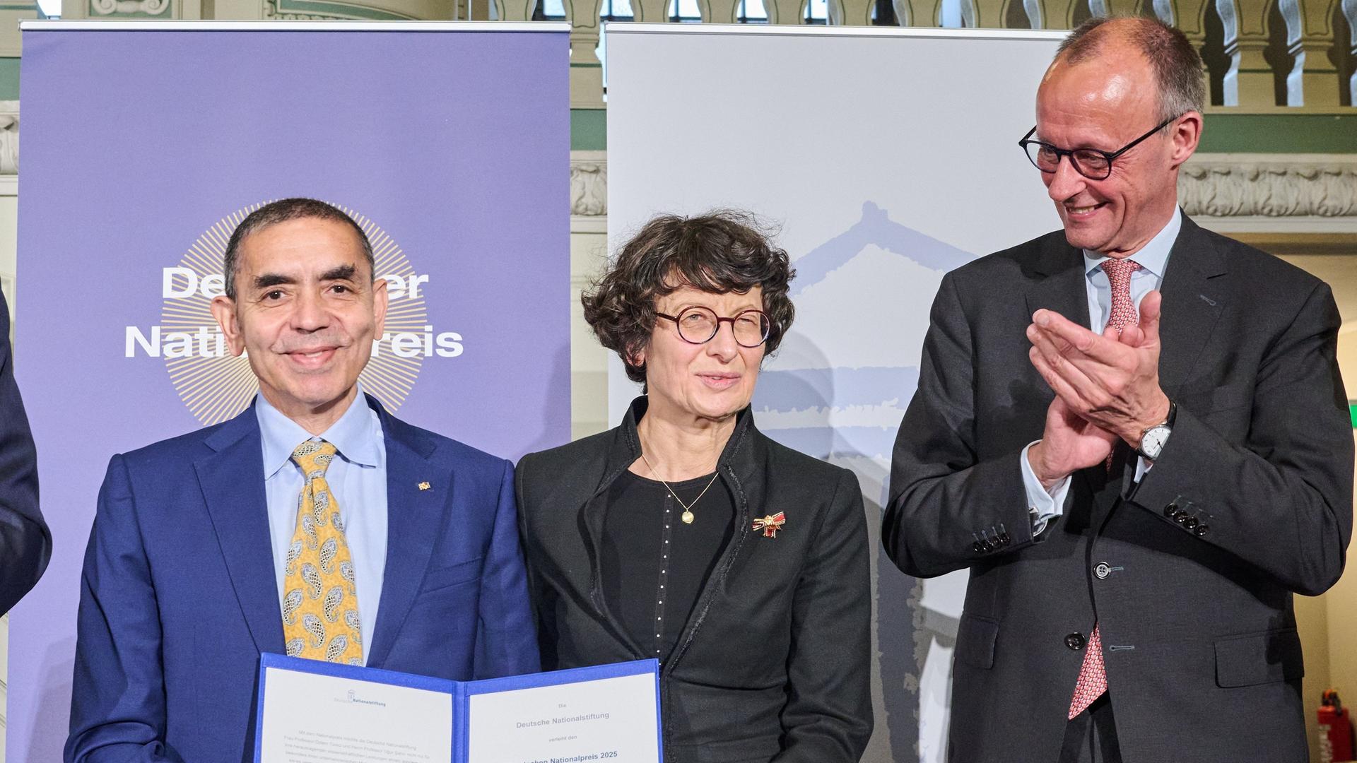 Das Professorenpaar, Gründer und Vorstandsvorsitzende des Unternehmens BioNTech Ugur Sahin (l-r) und Özlem Türeci, sowie Friedrich Merz (CDU), Bundeskanzler stehen bei der Verleihung des Deutschen Nationalpreises auf der Bühne im französischen Dom am Gendarmentmarkt.