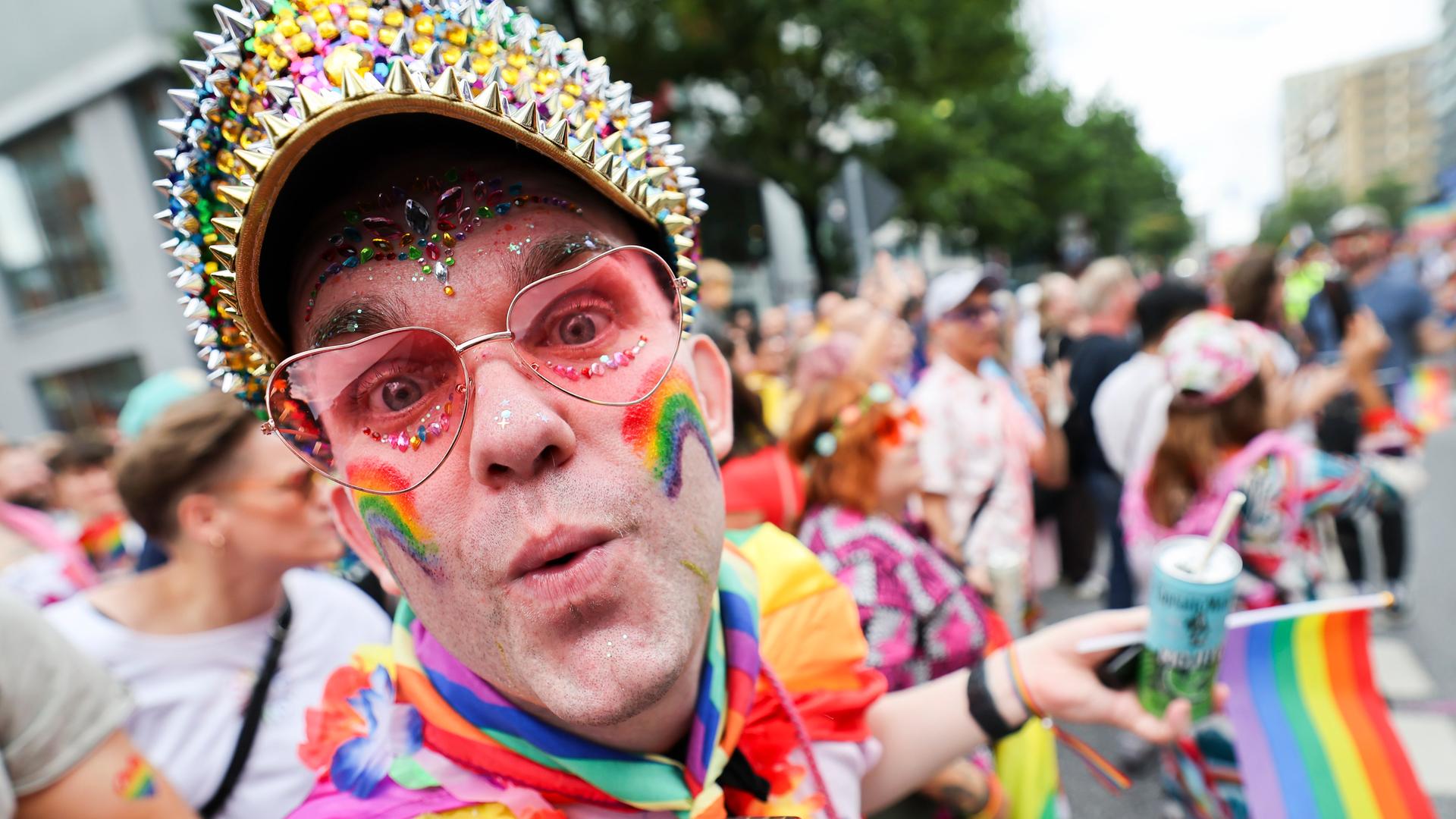 Nahaufnahme des Kopfes eines in Regenbogenfarben geschminkten Teilnehmers, der eine Mütze mit bunten Nieten trägt. Dahinter unscvharf der CSD-Umzug.