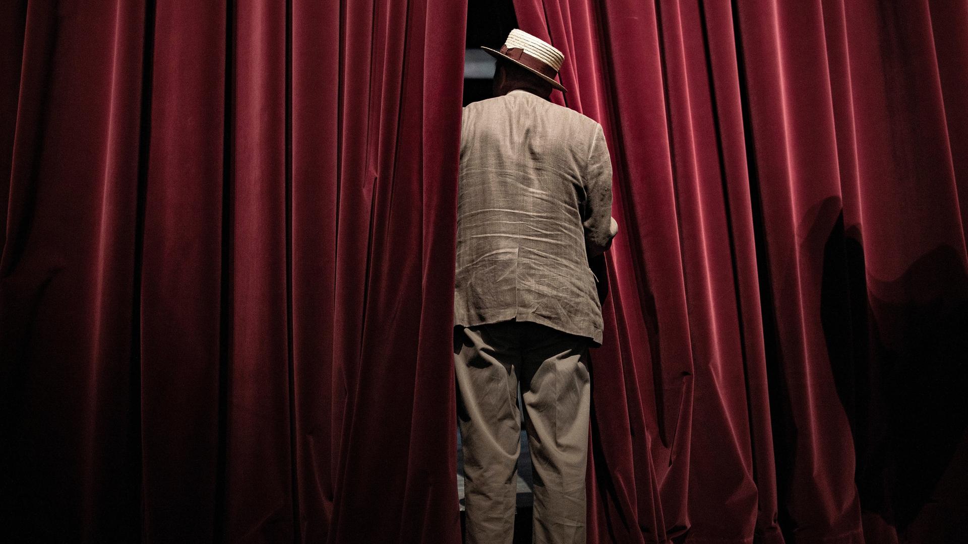 Ein Mann steht mit Anzug und Hut bekleidet in der Lücke eines roten Theater-Vorhangs, man sieht ihn von hinten