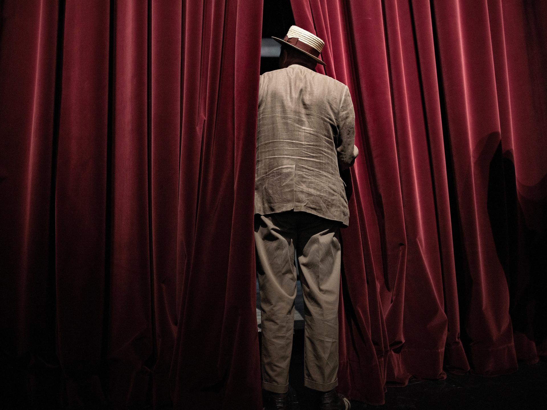 Ein Mann steht mit Anzug und Hut bekleidet in der Lücke eines roten Theater-Vorhangs, man sieht ihn von hinten