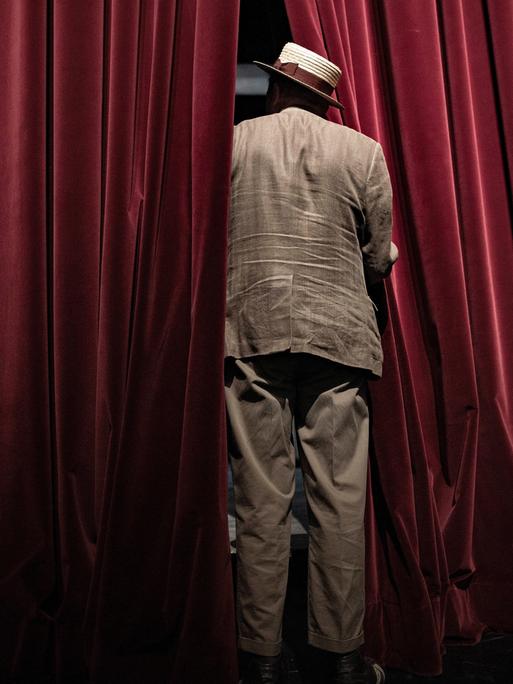 Ein Mann steht mit Anzug und Hut bekleidet in der Lücke eines roten Theater-Vorhangs, man sieht ihn von hinten
