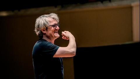 Ein Mann mit wilder, heller Mähne dirigiert in T-Shirt und lachend mit großer, lustvoller Geste.