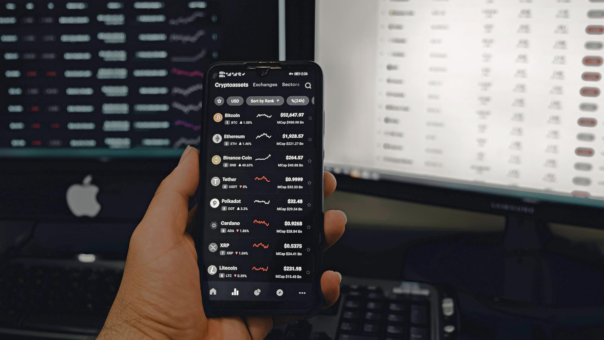 Eine Hand hält ein Smartphone, auf dessen Display mehrere Kryptowährungskurse zu sehen sind, im Hintergrund sind zwei Computer moniture zu sehen.
