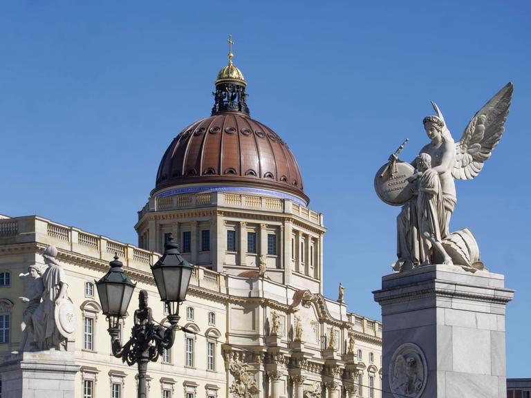 Die Kuppel des Berliner Stadtschlosses mit goldenem Kreuz. Im Vordergrund eine Skulptur zweier Engel.